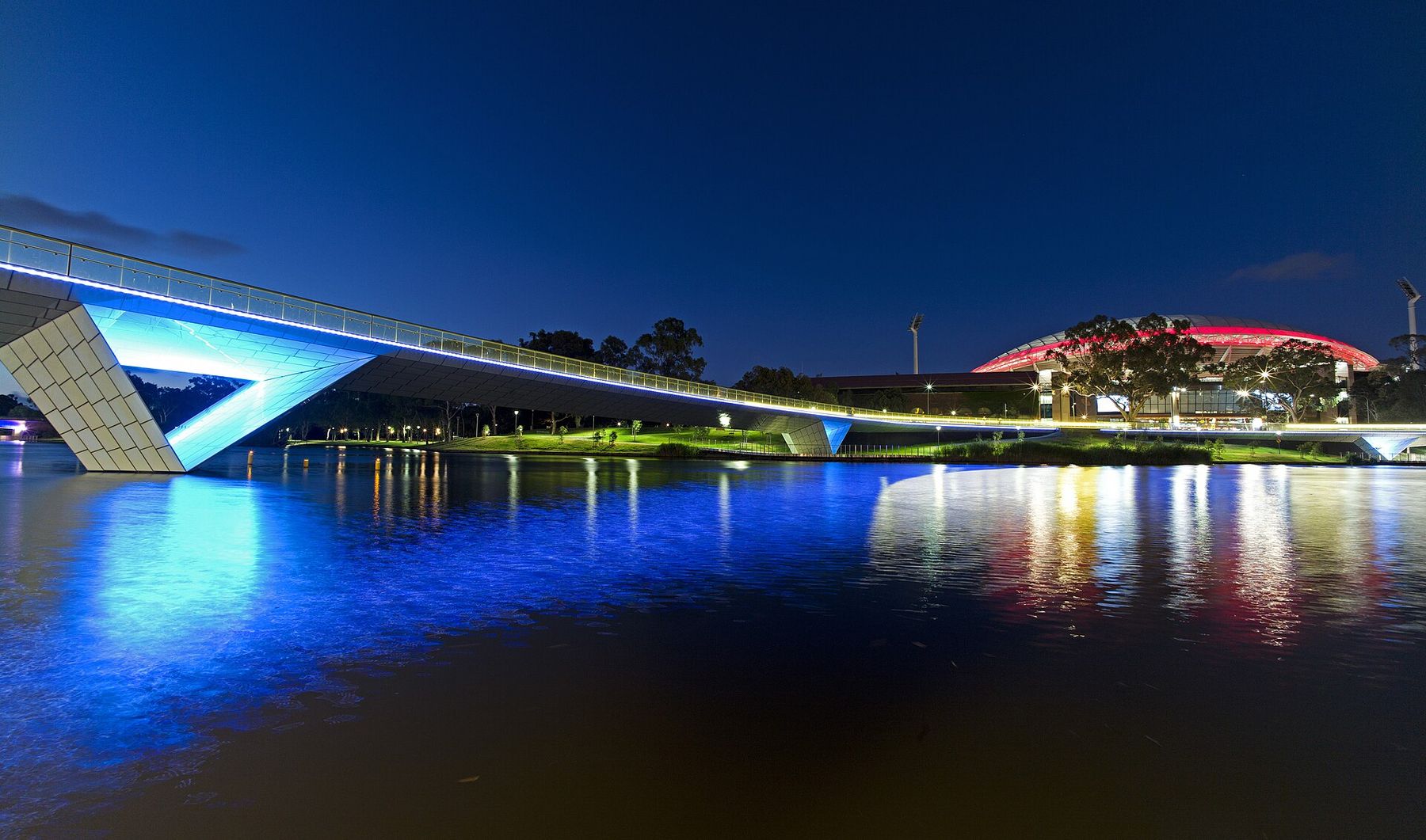 The illuminated Riverbank footbridge over the River Torrens at night, with Adelaide Oval glowing red behind